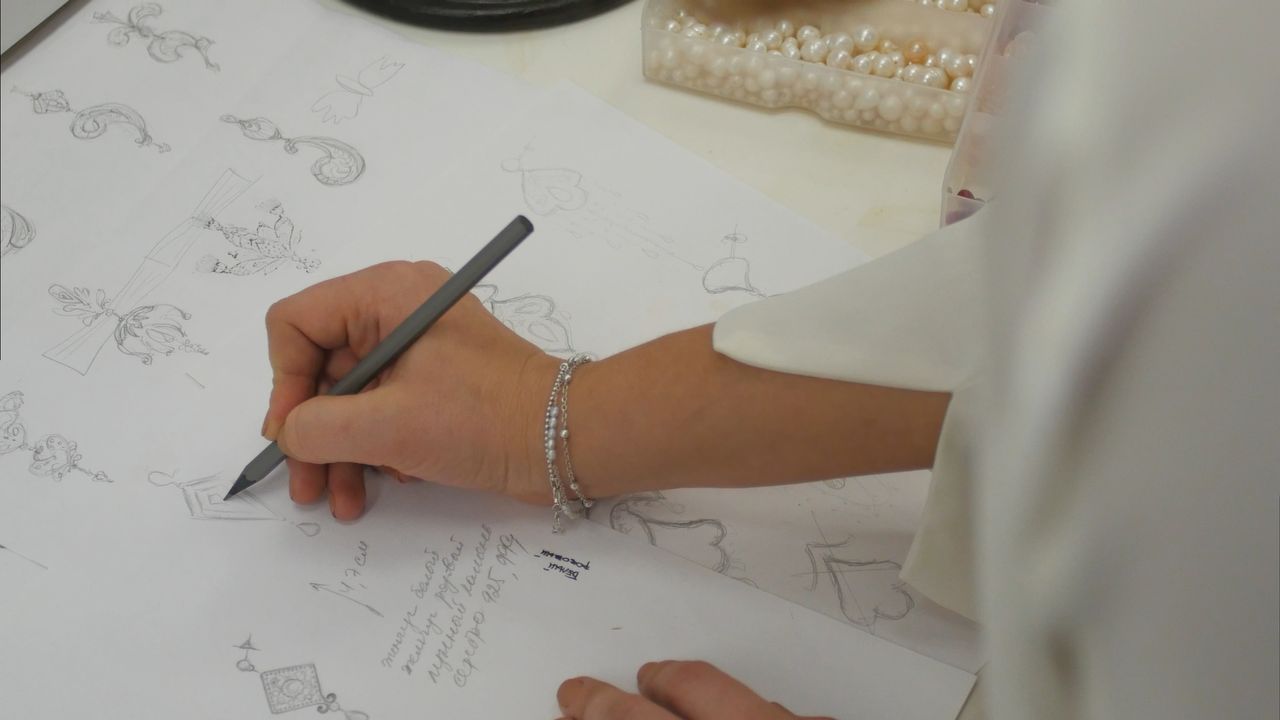 A woman drawing different jewelry designs on sheets of paper, a box of pearls next to her on the table.