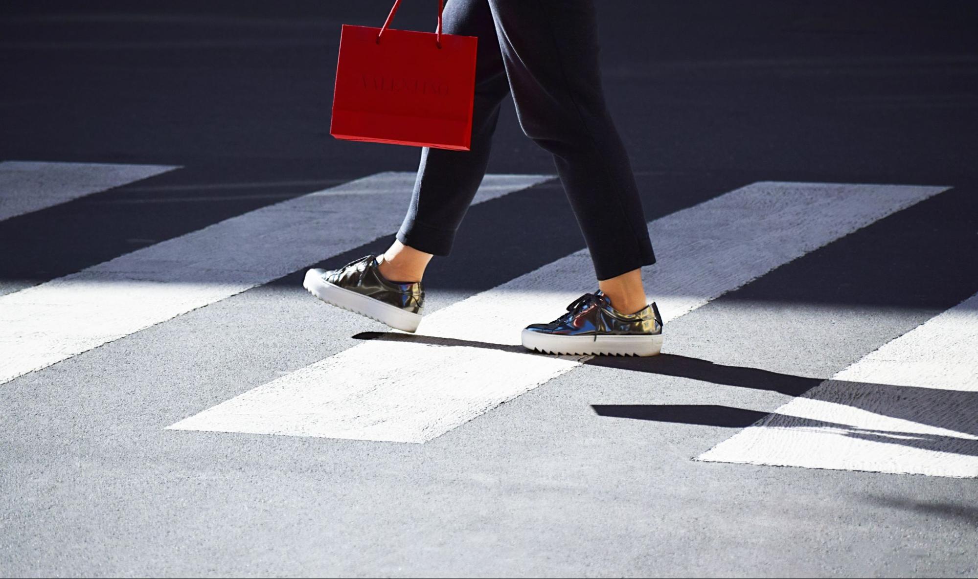 A close-up of feet walking on a crosswalk holding a red luxury shopping bag.