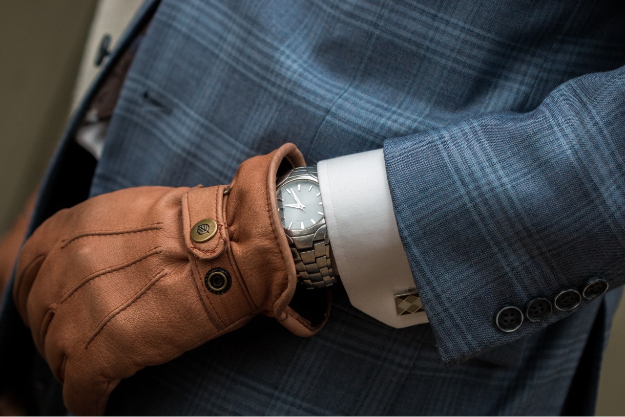 A close-up of a stainless steel watch on a man’s hand with leather glove on and a striped blue suit