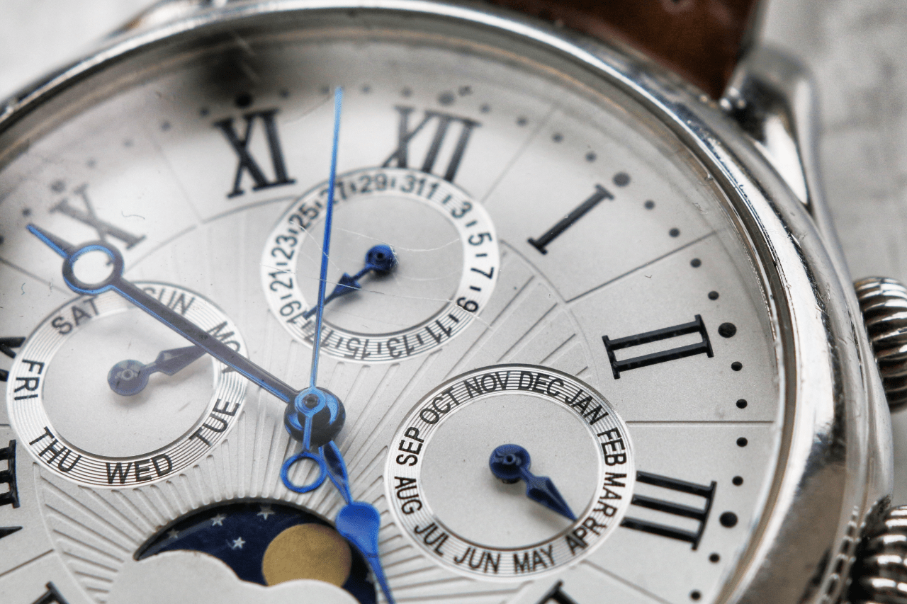 A close-up of a detailed watch white dial with roman numerals, deep blue hands and several subdials