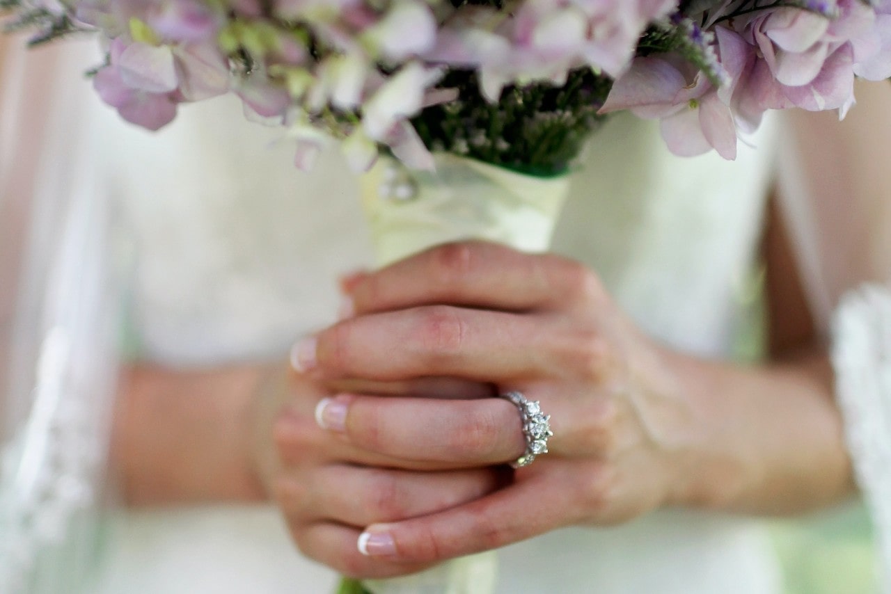 Close up image of a bride's hands holding a bouquet of flowers, wearing a three stone diamond engagement ring